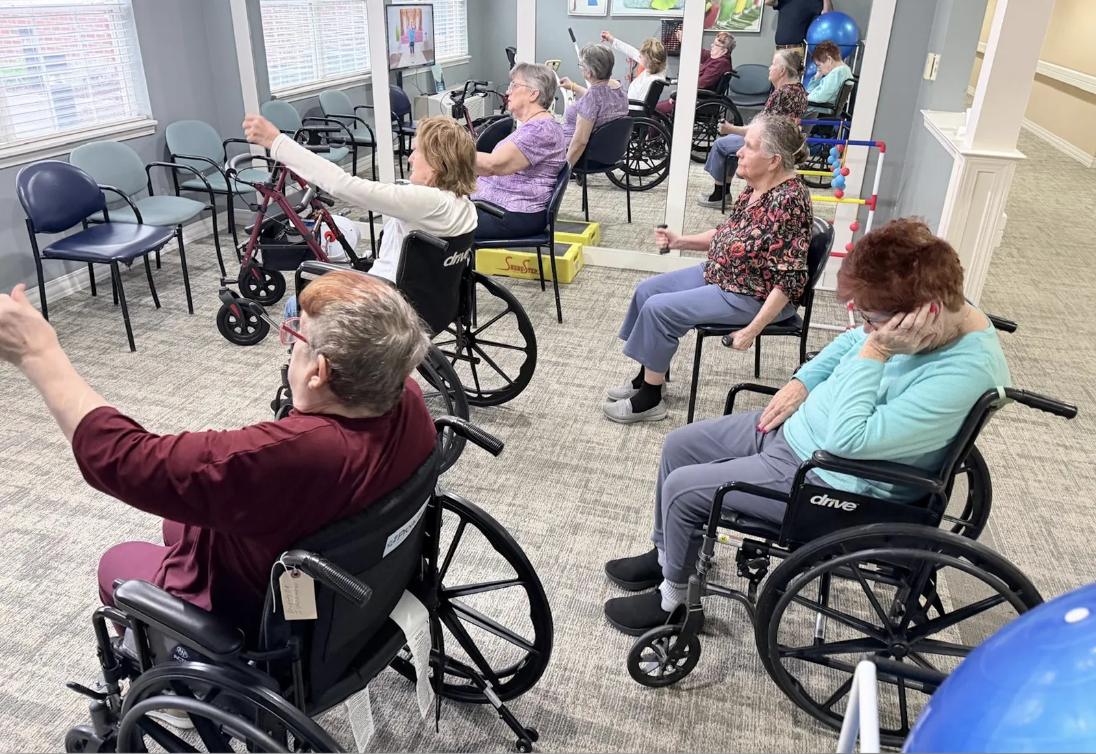 Residents in a seated CoveWell exercise class, arms raised, following the instructor on screen at Comfort Cove Senior Living in West Bloomfield, MI