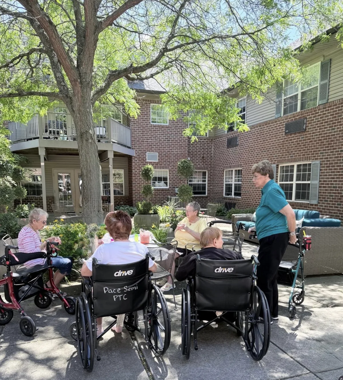 Residents gathered at a table in the shaded courtyard garden, with planters and patio seating against the brick building at Comfort Cove Senior Living in West Bloomfield, MI
