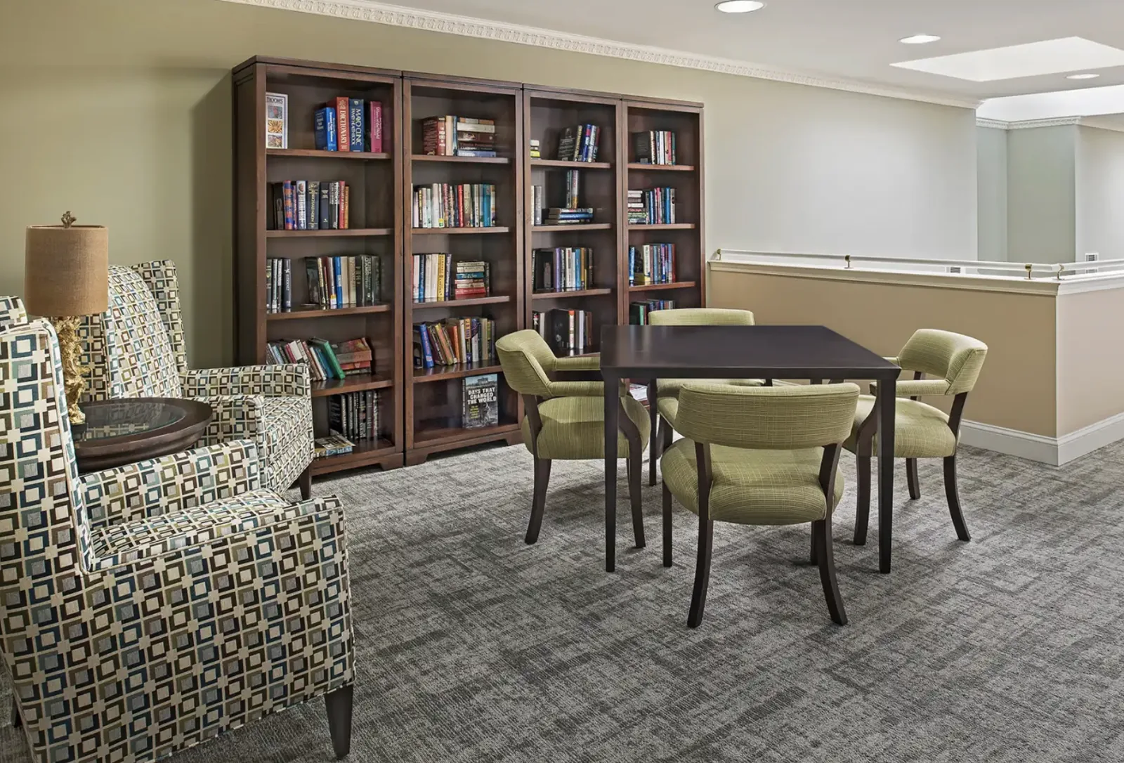 The library: bookcases, a games table with four chairs and two patterned armchairs at Comfort Cove Senior Living in West Bloomfield, MI