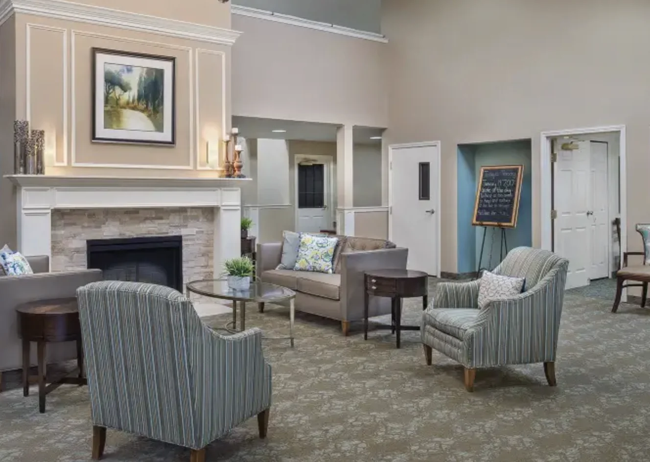 The lobby: striped armchairs and a sofa around a stone fireplace, with a chalkboard welcome sign by the doors at Comfort Cove Senior Living in West Bloomfield, MI