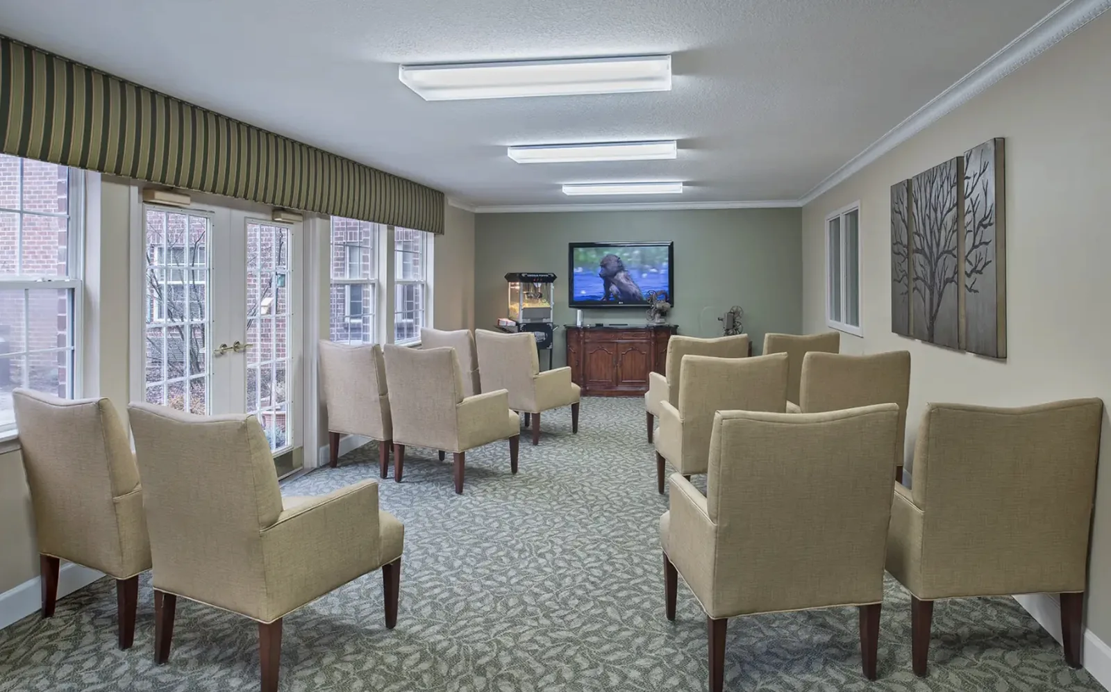 The movie theatre: rows of upholstered armchairs facing a wall-mounted screen, with a popcorn machine by the window at Comfort Cove Senior Living in West Bloomfield, MI