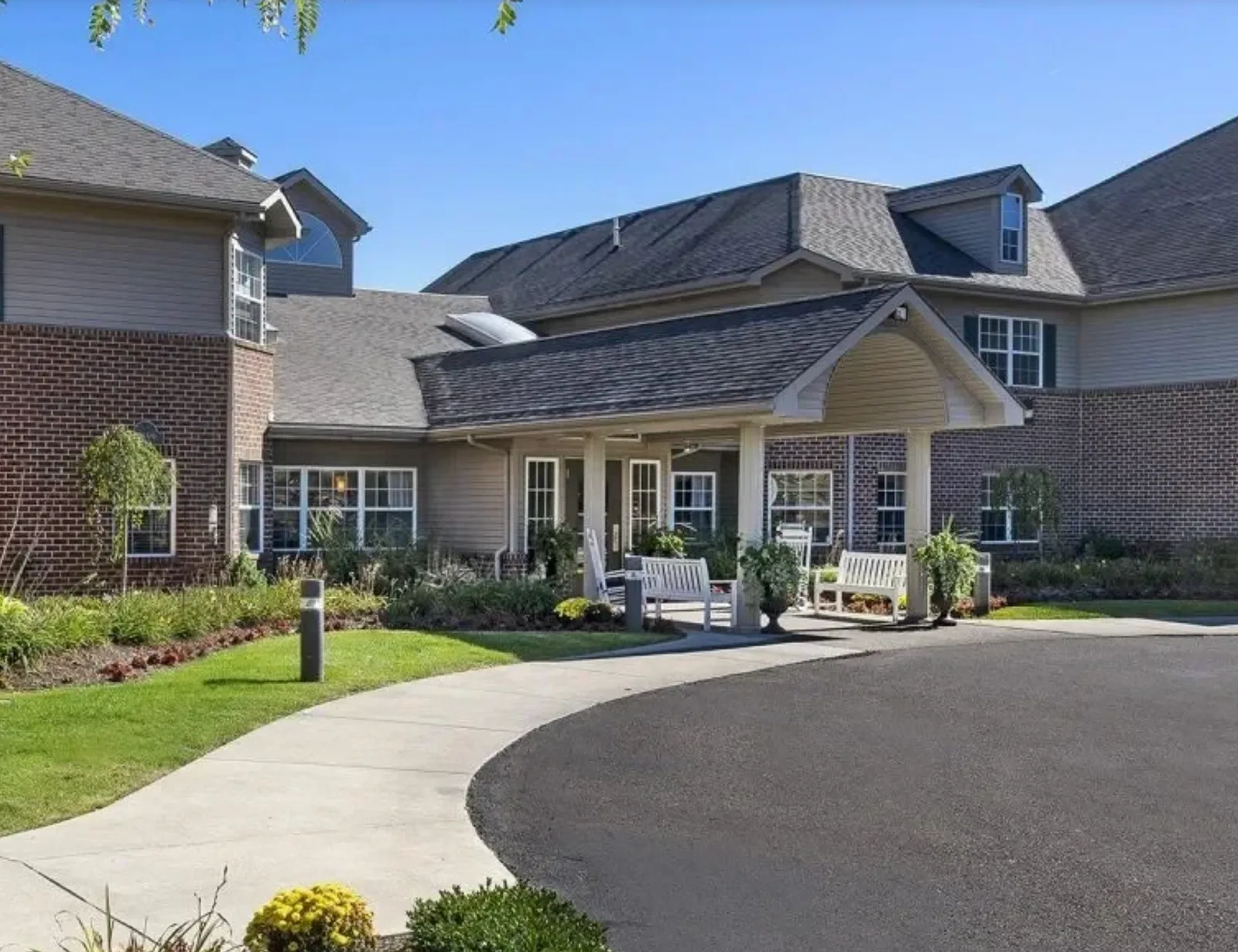 The entrance to Comfort Cove Senior Living in West Bloomfield, MI: brick and siding building with a covered portico, benches and rocking chairs, and the circular drive