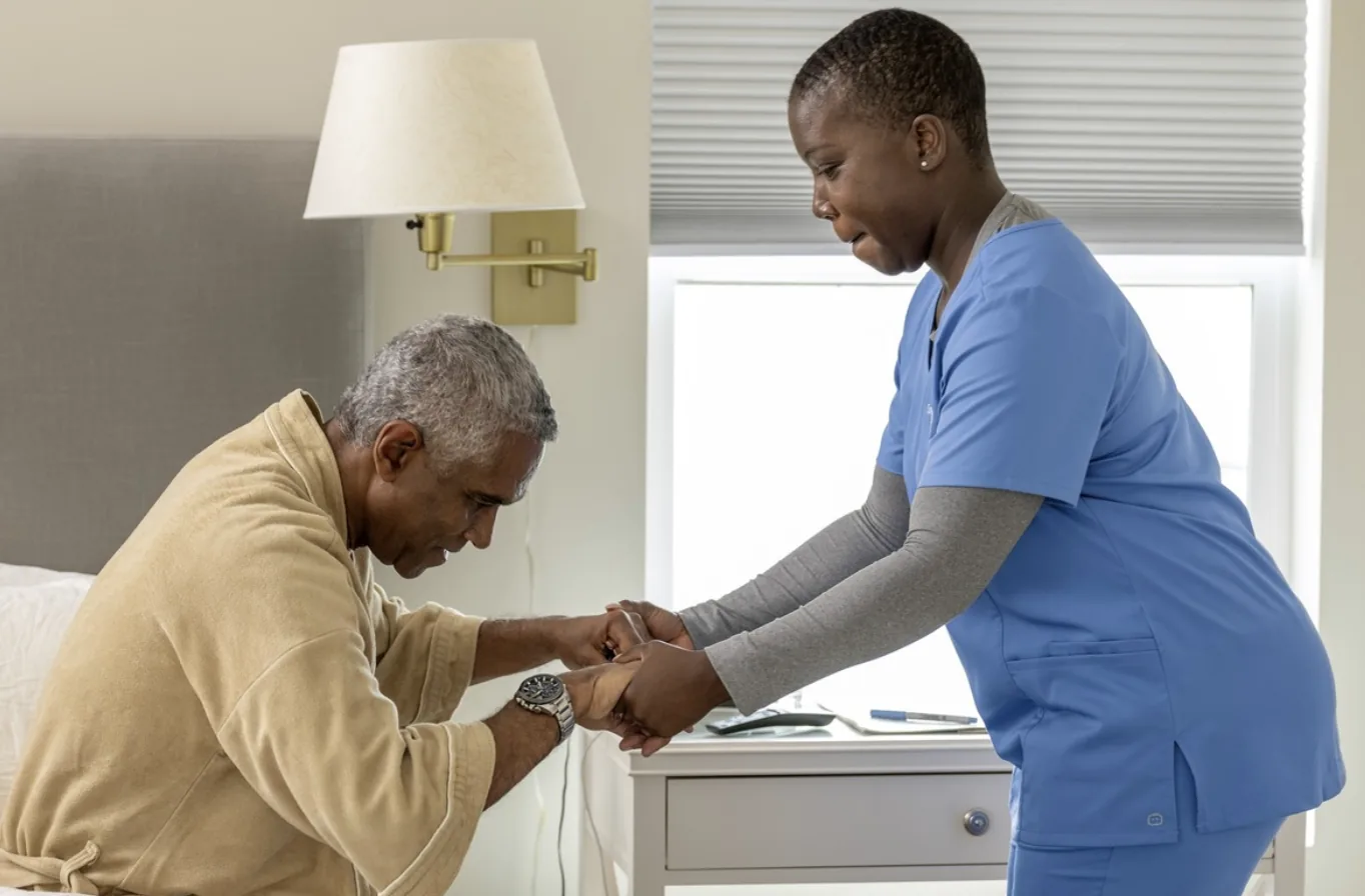 A ComForCare caregiver in blue scrubs helping a resident to his feet beside his bed at Comfort Cove Senior Living in West Bloomfield, MI